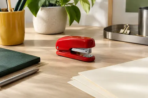 Agrafeuse de bureau compacte rouge près de feuilles ivoire et d’accessoires de bureau en métal mat.